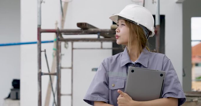 Asian female engineer walks slowly with clipboard near scaffold, performing construction site safety inspection, document checking, and quality control review inside unfinished building project.