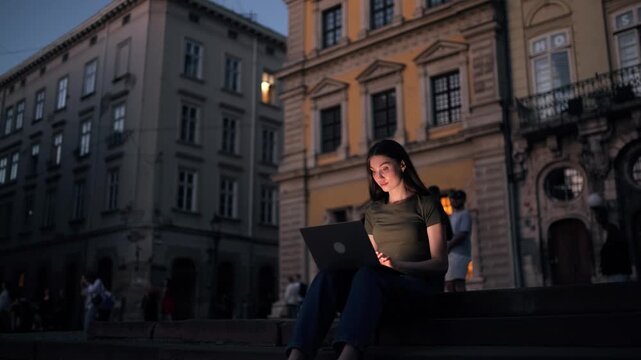 A young brunette woman sits on stone steps in an old European city square at night, focused on working on her laptop under warm street lighting