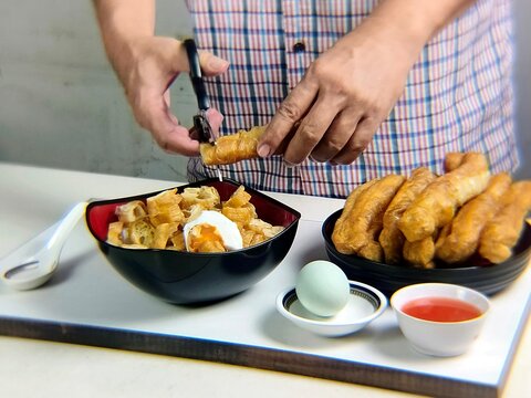 Man's hand cuts fried bread or cakwe using scissors over a bowl of porridge. Eating porridge topped with cakwe and salted egg is very popular among Chinese people as a breakfast dish.