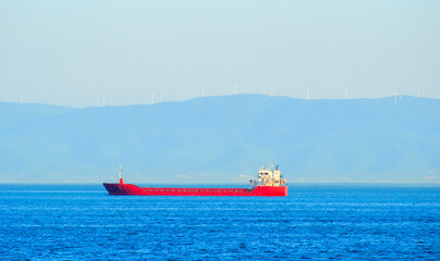 Fototapeta premium Red general cargo ship appearing small against the vast mountainous horizon