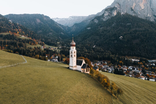 Aerial view of Chiesa di san Valentino with its white bell tower on a green hill surrounded by forests and mountains in Kastelruth, Trentino-Alto Adige, Italy.