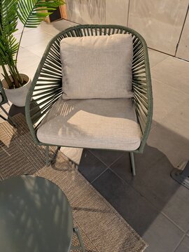 Rinc&oacute;n de una terraza con un sill&oacute;n de exterior con respaldo de cuerdas tensadas, una alfombra trenzada, una mesita redonda verde y una planta de hojas largas