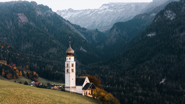 Aerial view of Chiesa di san Valentino on a grassy hill surrounded by pine forests and the Dolomite mountains Kastelruth, Trentino-Alto Adige S, Italy.