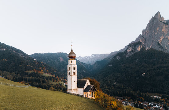 Aerial view of Chiesa di san Valentino on a grassy hill with the Dolomites mountains and autumn forests in the background in Kastelruth, Trentino-Alto Adige, Italy.