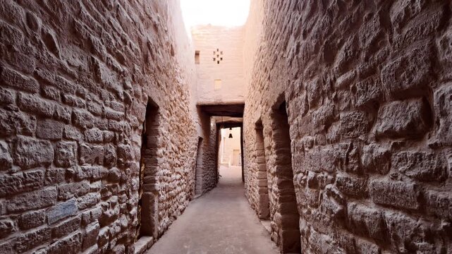 AlUla Old Town's narrow alley offering a view of ancient mud brick architecture and wooden doors under a bright blue desert sky, showcasing the historic heritage of Saudi Arabia