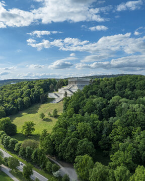 Aerial view of the Walhalla memorial on a lush green hillside surrounded by dense forest under a cloudy blue sky Donaustauf, Bayern, Germany.