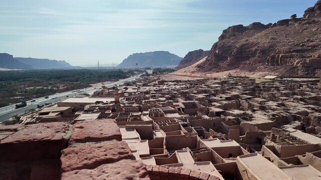 Wide aerial view across AlUla Old Town in AlUla, Saudi Arabia, highlighting the contrast between traditional mudbrick buildings, steep sandstone cliffs, and the historic fortification on the hillside.
