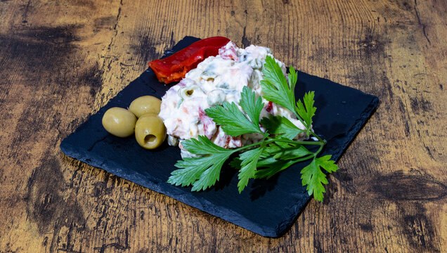 Ensaladilla Rusa Casera, con hoja de perejil, olivas rellenas y pimientos del piquillo sobre una plato de pizarra negro.