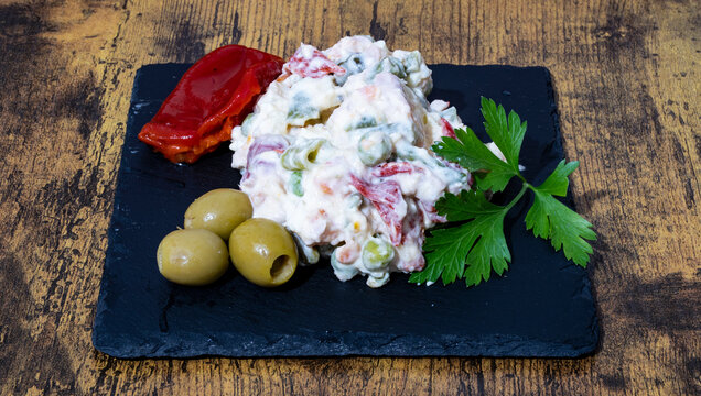 Ensaladilla Rusa Casera, con hoja de perejil, olivas rellenas y pimientos del piquillo sobre una plato de pizarra negro.