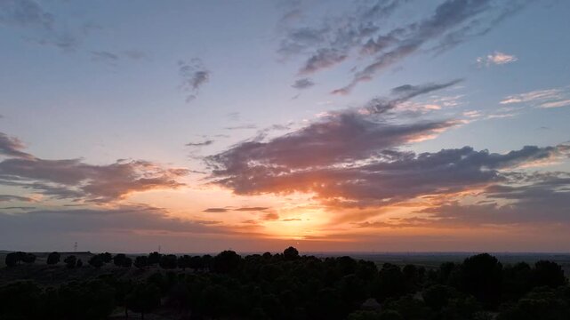 Aerial hyperlapse over Castilian plateau, Madrid, Spain. Sun sets on flat horizon silhouetting oak and pine trees against dramatic orange and golden sky with fast moving clouds.