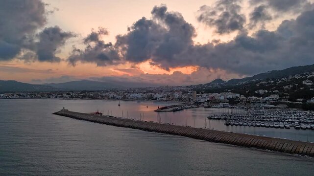 Aerial hyperlapse over Javea marina, Costa Blanca, Spain. Stone breakwater cuts diagonally across frame with sailboats and yachts under dramatic storm sunset reflected on calm water.
