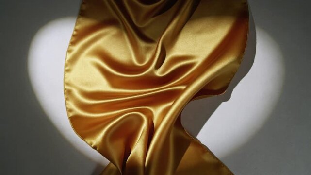 Top entering frame sending gold satin fabric falling to drape in studio under white heart spotlight