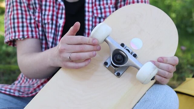 Girl spins wheels on skateboard checking their serviceability. Teenager preparing to ride, examining details of her first skateboard. Close-up of hands and skate outdoors. Skateboard inspection
