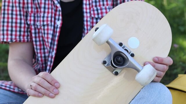 Girl spins wheels on skateboard checking their serviceability. Teenager preparing to ride, examining details of her first skateboard. Close-up of hands and skate outdoors. Skateboard inspection