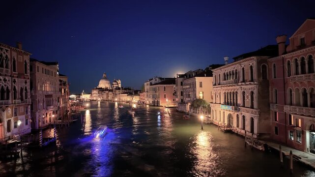 Night time-lapse video of the Grand Canal in Venice, Italy. Fast moving boats with light trails and illuminated Santa Maria della Salute basilica. Beautiful European cityscape and travel destination.