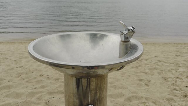 Outdoor Drinking Water Fountain Standing On Sandy Beach Near River. Infrastructure Object For Fresh Water Refreshment At Coastline
