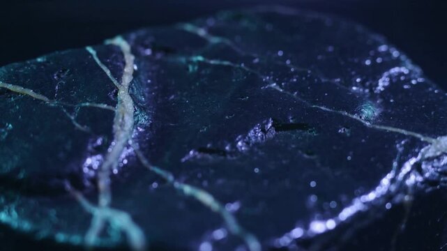 Shifting teal and purple lights revealing fissure and veins on round dark stone, accenting texture
