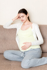 Obraz premium Young pregnant woman with headache sitting on sofa. Pregnancy expectation concept
