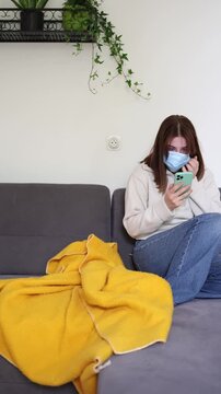 Vertical screen Young woman anxiously checks breaking news and messages on smartphone while wearing protective mask after rumors about another dangerous global virus begin spreading online