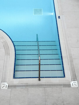 Modern indoor swimming pool entrance with stainless steel handrail, clear blue water, tiled steps, clean geometric design reflecting wellness, recreation, fitness lifestyle concept