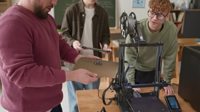 Teacher showing students plastic part, printed on flexible platform, while group examining result of open 3D printer with interest during school class