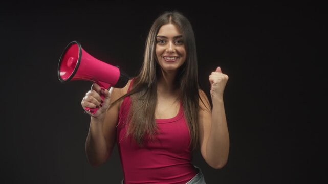 Woman holding pink megaphone with raised fist in studio, smiling in sleeveless tanktop; confidence empowerment activism.