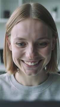 Young Caucasian woman with short blonde hair experiences a range of emotions while looking at a laptop screen in a bright indoor setting with shelves in the background