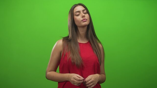 Young hispanic woman with hands clasping and slight head tilt, looking upward and fiddling fingers in a green screen studio; thoughtful reflection.