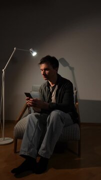 Vertical video man focusing on his smartphone screen in a dimly lit room, depicting concepts of loneliness, social isolation, technology addiction, and silent preoccupation