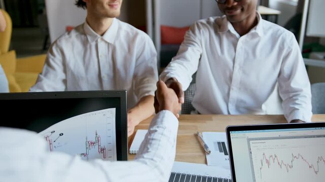 Two men shake hands in an office setting as they finalize a business deal while laptops display data charts and graphs in the background, capturing a moment of teamwork and agreement