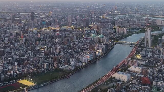 Aerial high-angle footage captures a panoramic evening view of Tokyo city from the Tokyo Skytree observation deck. The camera overlooks the winding Sumida River with its iconic bridges. 