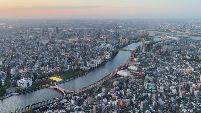 Aerial high-angle footage captures a panoramic evening view of Tokyo city from the Tokyo Skytree observation deck. The camera overlooks the winding Sumida River with its iconic bridges. 
