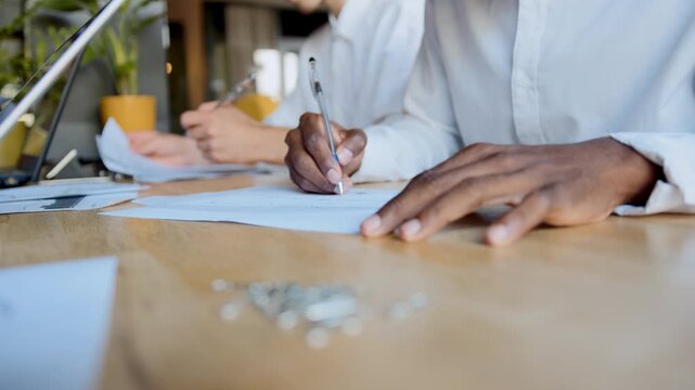 Black man writes on paper at a desk while another person is focused on a task in a workspace during the daytime in a modern office setup