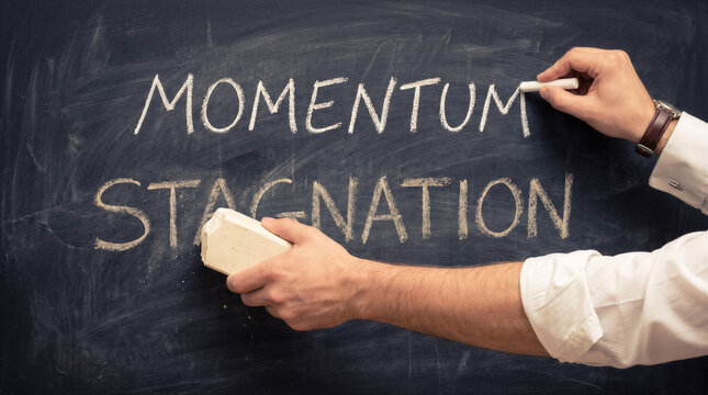 Closeup of a person writing the word momentum on a chalkboard while simultaneously erasing the word stagnation with an eraser