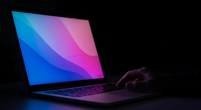 Close-up of glowing laptop screen with abstract colors and a hand typing on the keyboard in a dark environment