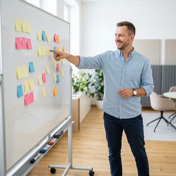 Smiling caucasian male marketing specialist in his 30s in a casual blue shirt pointing at a whiteboard covered with colorful sticky notes in a bright modern office