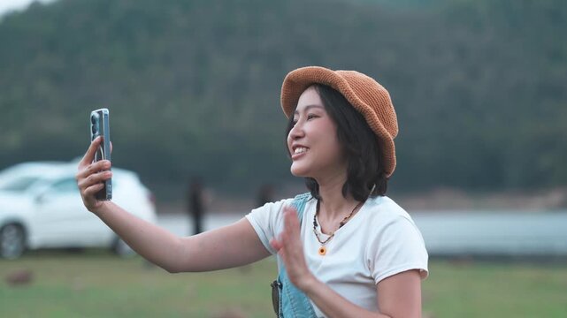 Happy young Asian woman traveler holding a smartphone and waving her hand while making a video call or recording a vlog at an outdoor campsite