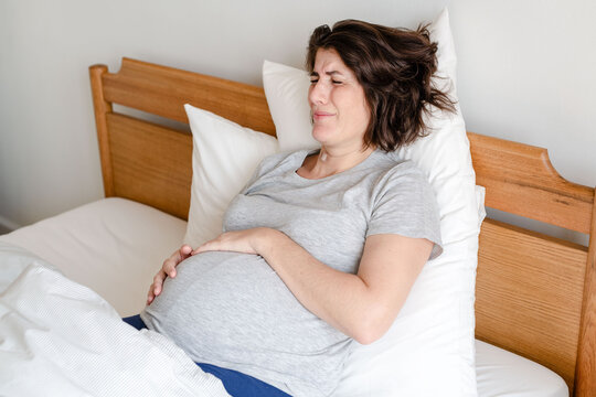 Pregnant woman in discomfort lying on a bed, holding her belly.