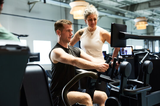 Male client receives personal training on rowing machine from coach in modern gym. 