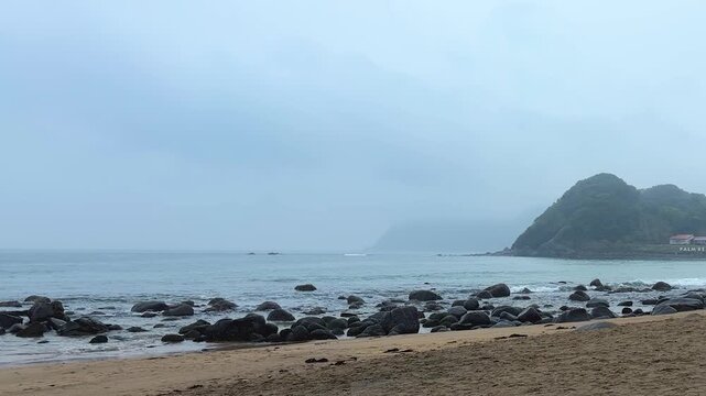 A gloomy view opens up over the sandy and rocky beach in Itoshima, Fukuoka. Dark rocks stretch out into the ocean under a thick blanket of grey clouds. 