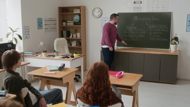 Handheld zoom in shot of male teacher giving lecture on 3D modelling basics, using chalkboard to group of attentive middle school students