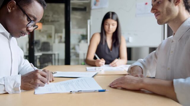Professional hands sign documents at a table. Two men shake hands, completing a deal. Woman observes the agreement closely. Business success, collaboration, and trust are key.