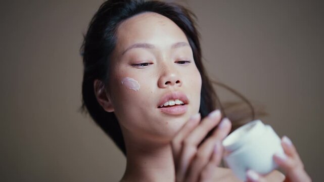 Young asian woman with freckles and long straight black hair, gently touching her face and smiling, expressing happiness and confidence on a neutral background	