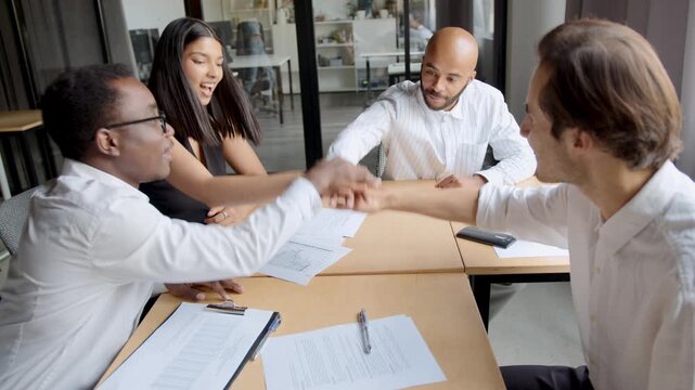 Four diverse business people, two men and two women, stack their hands together on a meeting table in an office, symbolizing teamwork and agreement