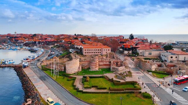 Aerial view of ancient fortress ruins in historic Nessebar town. High angle view of historic stone fortress walls, green lawns, harbor with boats, and red roofs under cloudy sky.