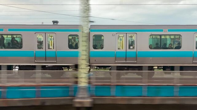 A passenger train passes over a steel truss bridge in Japan. The commuter train moves steadily on a multi-track railway line. High angle view shows suburban infrastructure with green riverbanks.