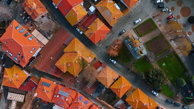 Top down aerial view of old town street intersection layout. Direct vertical view of terracotta tiled roofs, stone paved streets, parked cars and a small green park.