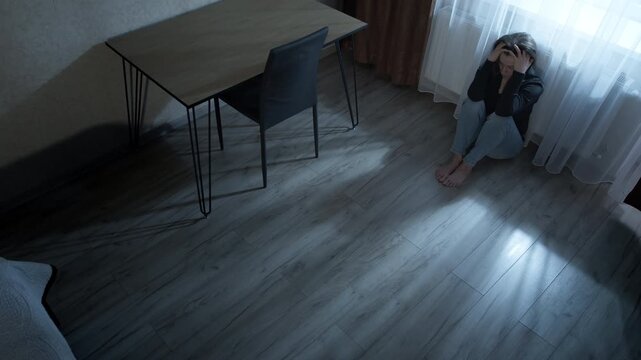 Sad lonely woman sitting on the floor in dark room near window suffering from depression. Desperate female crying alone inside house representing psychological pain and mental health concept.