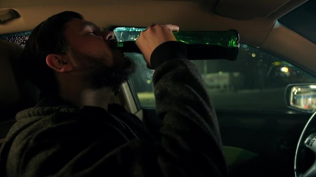 Side view of bearded caucasian man drinks alcohol inside a car at night. Young person with closed eyes in vehicle regrets drinking whiskey from a green bottle. Violation of the sobriety driving law