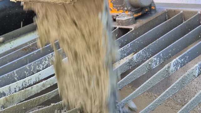 Extreme close up diagonal rear view of ready mix concrete pouring from concrete mixer truck into the hopper of a concrete pump at a construction site in normal speed